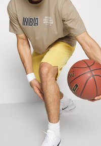 Camiseta con el logo de la NBA en caqui, pantalones cortos amarillos, calcetines deportivos blancos y zapatillas blancas. Sosteniendo un balón de baloncesto marrón en una postura baja.
