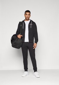 Sweat à capuche noir zippé avec cordons blancs, associé à un pantalon de jogging noir et des baskets blanches. Tient un sac de sport noir avec des accents en maille.