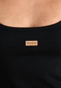 Must mustriba tank-top, mille laia kaelusega, millel on tan värvi ristkülikukujuline silt, mille keskel on "HUGO". Sile tekstuur.