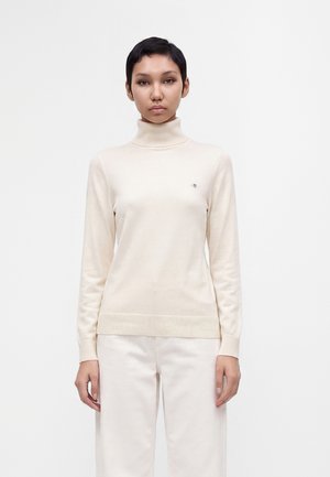 Femme aux cheveux courts et foncés, portant un pull à col roulé crème et un pantalon assorti, debout devant un fond clair uni.