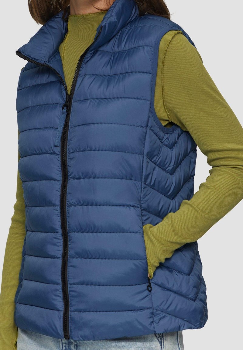 Femme portant une veste sans manches matelassée bleue par-dessus une chemise à manches longues vert olive, avec une main dans la poche de la veste.