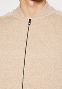 Giacca bomber color beige chiaro in tessuto texturizzato, con colletto a costine e una cerniera nera sul davanti. Presenta dettagli hardware minimi.