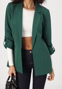 Zielona marynarka o dopasowanym kroju z miękką fakturą, z pojedynczym zapięciem na guzik i podwiniętymi mankietami. Noszona na białej bluzce i ciemnych dżinsach.