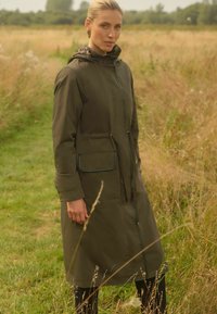 Olivově zelený nepromokavý kabát s kapucí na šňůrku, dvěma předními kapsami a kontrastním lemováním. Hladká textura, délka do kolen, volný střih.