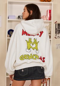 Bela majica s kapuco z ohlapnim krojem, na kateri so pisani grafični motivi s temo Grincha: "Vesele Grinchmas" v odebeljeni rdeči in rumeni barvi, z zelenim dizajnom rok.
