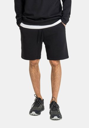 Mann trägt schwarze Strickshorts, schwarzen Pullover über weißem Hemd und schwarze Turnschuhe mit gemusterten Schnürsenkeln, steht mit Händen in den Taschen.