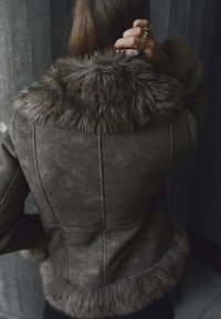 Persona con cabello largo vestida con una chaqueta de piel de oveja marrón con cuello y puños de piel, mano levantada hacia la cabeza, de espaldas a la cámara.