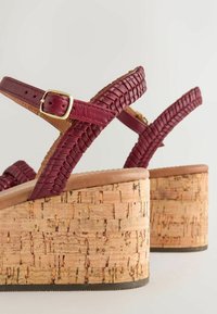 Sandales compensées avec talons en liège texturé et brides en cuir rouge foncé tressé avec fermeture à boucle réglable.