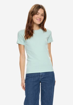 Kvinde smilende, iført lyseblå kortærmet skjorte og blå jeans, stående mod en ensfarvet hvid baggrund.