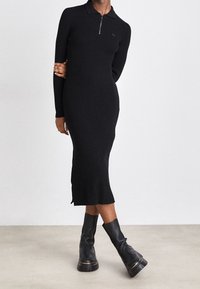 Robe noire en tricot côtelé avec un col zippé, coupe slim, longueur genou, fentes latérales, portée avec des bottes noires montantes à semelles épaisses.