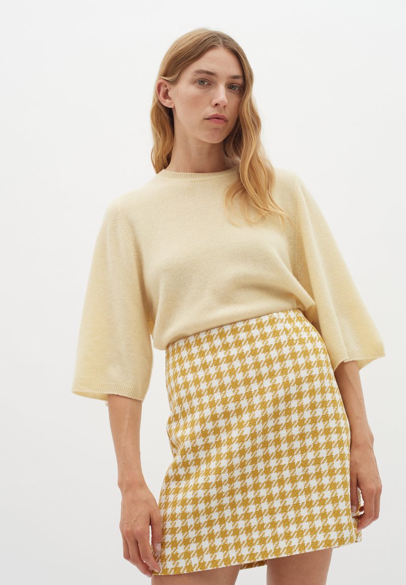 Kreminės spalvos megztinis su alkūnių ilgio rankovėmis, derinamas su geltonai ir baltai dryžuota mini sijonėliu.