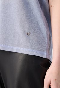 Gros plan d'une personne portant un pull tricoté bleu clair avec un petit logo métallique rond et un pantalon en cuir noir.