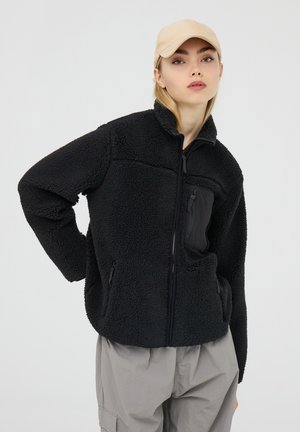 Giacca in pile nera con colletto alto, tasche con zip e finitura texturizzata. Abbinata a pantaloni grigio chiaro e a un cappello beige.