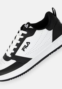 Zapatilla blanca y negra con parte superior de cuero, detalles perforados, cordones planos blancos y suela de goma negra. Logo de Fila en el lateral.