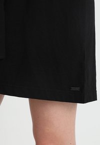 Gros plan d’un ourlet de jupe noire au-dessus du genou avec une petite étiquette rectangulaire de marque près du bord inférieur sur un fond de peau nue.