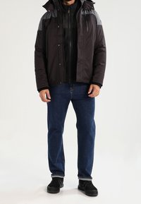 Schwarz-graue, isolierte Jacke mit Kapuze, ausgestattet mit einer Reißverschlusstasche auf der Brust, kombiniert mit blauen Jeans und schwarzen Sneakers.