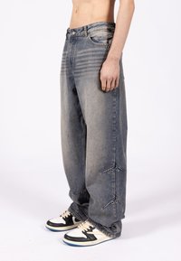 Φαρδιά μπλε τζιν με ξεθωριασμένο φινίρισμα, με οριζόντιες ρίγες, μοναδικά ράμματα και λεπτομέρειες με μεταλλικά κρικάκια στο τελείωμα. Συνδυασμένα με αθλητικά παπούτσια.