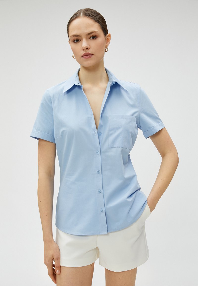Camisa de manga corta de color azul claro con cuello, cierre frontal de botones y un bolsillo en el pecho izquierdo. Confeccionada en una tela suave y con un corte ligeramente ajustado.