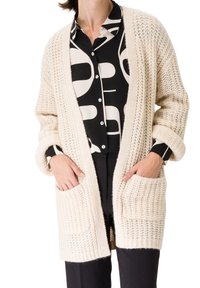 Personne portant un cardigan en maille épaisse crème avec des manches retroussées, par-dessus une chemise boutonnée à motif abstrait noir et blanc et un pantalon noir.