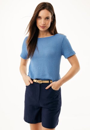 Frau mit langen braunen Haaren, die ein hellblaues kurzärmeliges Strickoberteil und marineblaue Shorts mit einem beigen Gürtel trägt und mit den Händen in den Taschen steht.