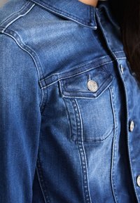 Denim jacket in medium blue, featuring a collar, two chest pockets, silver button accents, and a smooth texture with subtle fading.
