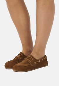 Zapatos náuticos de ante marrón con diseño de cordones, ojales de tono dorado, textura suave y puntera redonda. Suela parece flexible y plana.