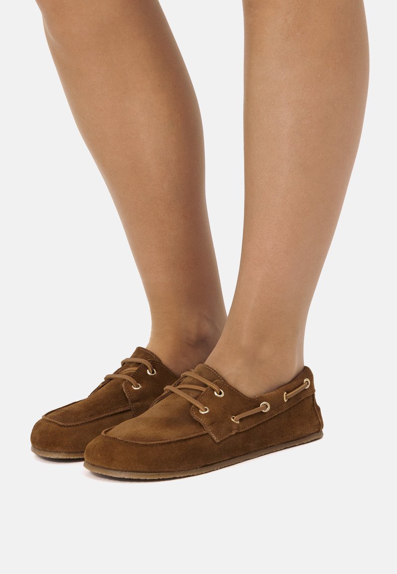 Zapatos náuticos de ante marrón con diseño de cordones, ojales de tono dorado, textura suave y puntera redonda. Suela parece flexible y plana.