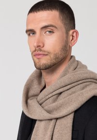 Sciarpa di lana beige drappeggiata attorno al collo, caratterizzata da una consistenza morbida e una finitura liscia e uniforme, che completa un cappotto nero.