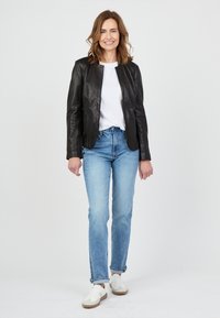 Zwarte leren jas met een kraag, gecombineerd met een witte t-shirt en lichtblauwe jeans. Model draagt witte sneakers met beige accenten.