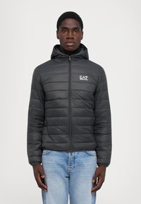 Homme portant une veste à capuche matelassée noire avec le logo EA7 Emporio Armani, se tenant contre un fond blanc uni, les mains sur les côtés.