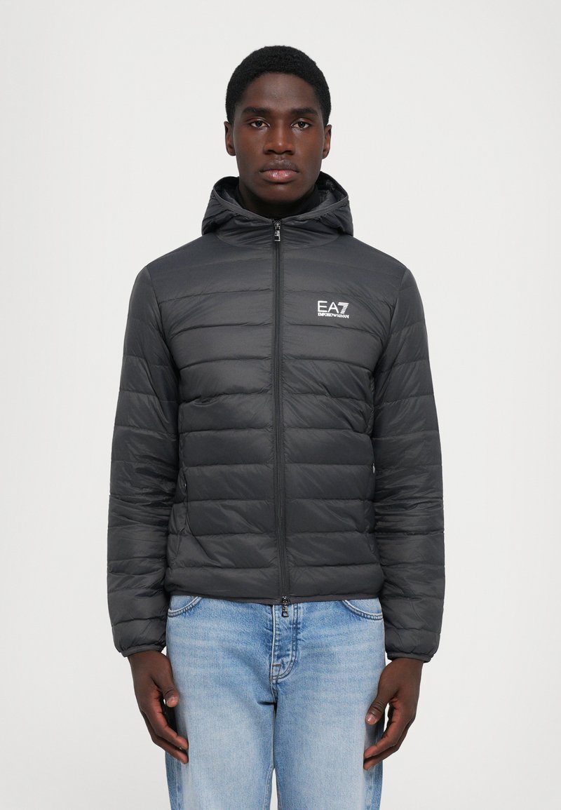 Homme portant une veste à capuche matelassée noire avec le logo EA7 Emporio Armani, se tenant contre un fond blanc uni, les mains sur les côtés.