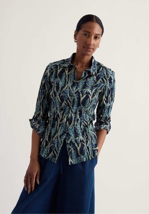 Femme portant une chemise ajustée à manches longues avec des motifs de feuilles bleues et vertes et un pantalon large bleu foncé, debout devant un fond uni.