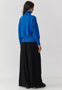 Niebieski sweter z golfem, wykonany na szydełku, w luźnym fasonie, z prążkowanymi mankietami i dołem, w zestawieniu z czarnymi spodniami o szerokiej nogawce, widok z tyłu.