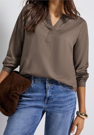 Langærmet, olivengrøn bluse med V-hals og rynkede manchetter, kombineret med blå denimbukser. Tilbehøret inkluderer en brun ruskindsklæder.