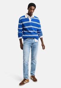 Jeune homme portant un pull rayé bleu et blanc par-dessus une chemise blanche, un jean bleu clair et des baskets marron, se tenant devant un fond blanc.