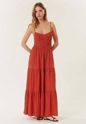 Jeune femme portant une robe maxi sans manches couleur rouille à volants avec fines bretelles et sandales noires, debout devant un fond neutre.