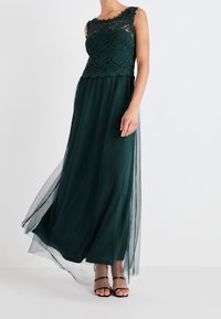 Lång grön klänning med spetsliv, ärmlös design och flödande genomskinlig kjol. Sandaler med vadderade remmar kompletterar looken.