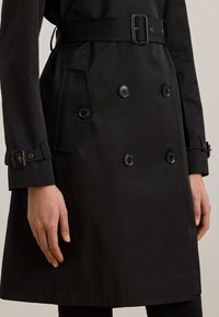 Musta kaksirivinen takki, jossa vyö ja napit kalvosimissa ja edessä, henkilö pukeutuneena, näkyvissä yksi käsi sivulla.