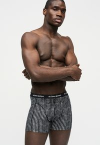 Μυώδης άνδρας στέκεται με τα χέρια σταυρωμένα, φορώντας μαύρα και λευκά μποξεράκια με σχέδιο Björn Borg, μπροστά σε απλό ανοιχτό φόντο.