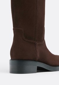 Smeđi suedni čizme do koljena s teksturiranim završetkom, naglašene čvrstim crnim blok petama i čistim detaljima šivanja duž bočnih strana.