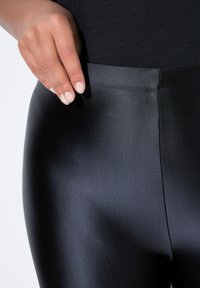 Zwarte leggings met een gladde, glanzende textuur. De tailleband is vlak en naadloos. Een hand houdt lichtjes de bovenrand van de leggings vast.