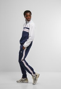 Marineblå og hvit treningsoverall med fargeblokkerende design og røde striper, jakke med glidelås, elastiske mansjetter og tettsittende joggebukser.