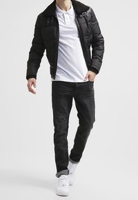 Chaqueta acolchada negra con cuello de piel, camiseta polo blanca, vaqueros oscuros y zapatillas blancas, con cierre de cremallera y detalles de costuras sutiles.