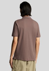 Camisa de polo marrón de manga corta hecha de una tela suave, con un cuello estándar y un corte recto, combinada con pantalones de color claro.