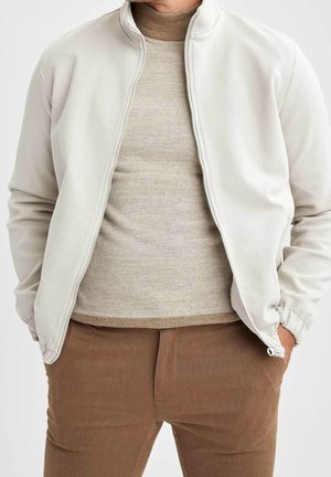 Mand iført beige jakke med lynlås over lys brun tekstureret sweater og brune bukser, hænderne i lommerne, overkrop synlig.