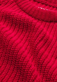 Gros plan d'un tissu tricoté rouge vif avec une texture côtelée et un bord de col plié, montrant des motifs de points détaillés.