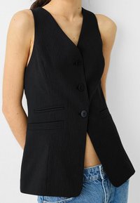 Svart kostymväst med V-ringning, fem knappar och två sidofickor. Tyget har en slät yta, som kombineras med högmidjade denimshorts.