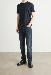 Man i mörk marinblå t-shirt, svart bälte, mörkblå jeans och svarta skor som står på ett grått golv mot en vit vägg.