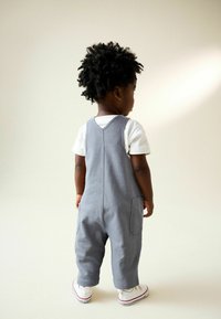 Copil mic cu păr creț, îmbrăcat în salopetă gri, bluză albă și adidași albi, stă cu spatele la fundal simplu.