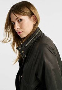 Czarna kurtka zapinana na zamek z złotymi akcentami, z wysokim kołnierzem i detalami na ramionach. Wykonana z gładkiego, lekkiego materiału o matowym wykończeniu.